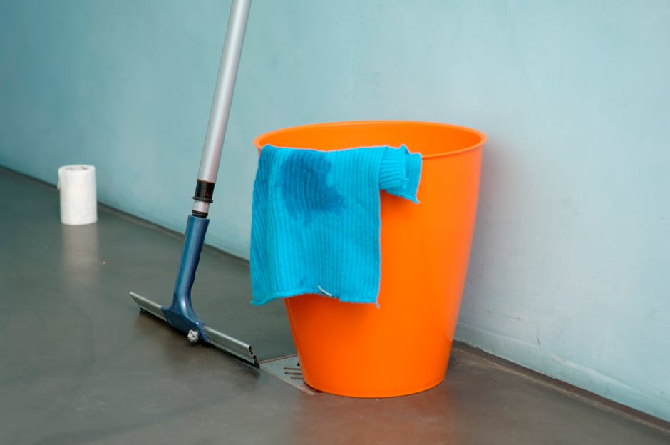 Arrangement of cleaning tools and supplies on a white surface, including a pair of pink rubber gloves, a pink cleaning cloth, a green spray bottle, a yellow and red sponge, and a pale pink bottle of c