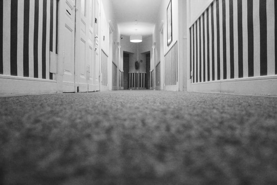 A person wearing casual clothing and white sneakers is operating a vacuum cleaner on a patterned beige and green carpet inside a room. The vacuum cleaner has a yellow body with a black hose attachment