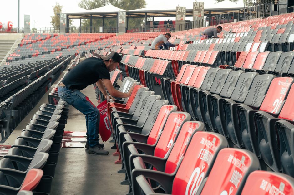 Two workers are cleaning the seats and aisles of an outdoor stadium with tiered red and gray plastic seats, in preparation for an event. The worker in the foreground is wiping a red seat with a cloth, while the other two are cleaning the seats in the background. The stadium appears well-maintained and is equipped with black armrests and designated seating sections, with natural daylight illuminating the scene. The image emphasizes surface cleaning and maintenance, aligning with professional deep cleaning and sanitisation services provided by Carpet Cleaners Brent for commercial venues such as Wembley Stadium, reflecting hygiene and cleanliness standards for large-scale public spaces.
