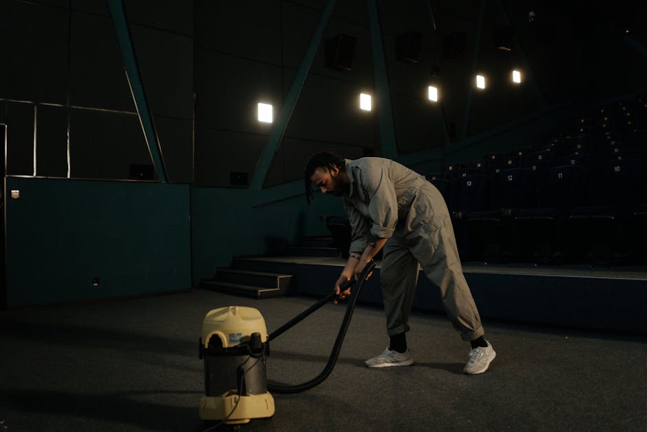 A professional cleaner from Carpet Cleaners Brent is performing deep surface cleaning in the interior of Wembley Stadium. The individual is wearing light-colored overalls and white sneakers, using a vacuum and cleaning tools on a dark, carpeted floor. The room features tiered seating with dark blue upholstery, a modern architectural design with angled wall panels, and several bright ceiling lights providing illumination. The area appears spotless and well-maintained, highlighting the company's expertise in sanitisation and domestic cleaning services related to large-scale venues.